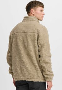 Beiger Fleece-Pullover mit hohem Kragen, strukturiert, leicht überschnittener Passform und sichtbaren Nähten auf dem Rücken und den Ärmeln.