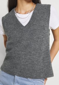 Gilet grigio a maglia con scollatura a V, caratterizzato da una texture a coste. Indossato sopra una maglietta bianca, l'orlo è leggermente più lungo sul retro.