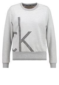 Sudadera gris de manga larga, con cuello redondo y letras negras con tachuelas "CK" en el frontal. Tejido suave con dobladillos y puños acanalados.