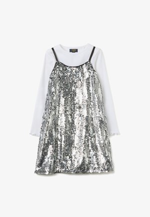 Vestido plateado con lentejuelas y tirantes oscuros ajustables, superpuesto sobre una camisa blanca de manga larga con puños con volantes, creando un efecto reflectante.