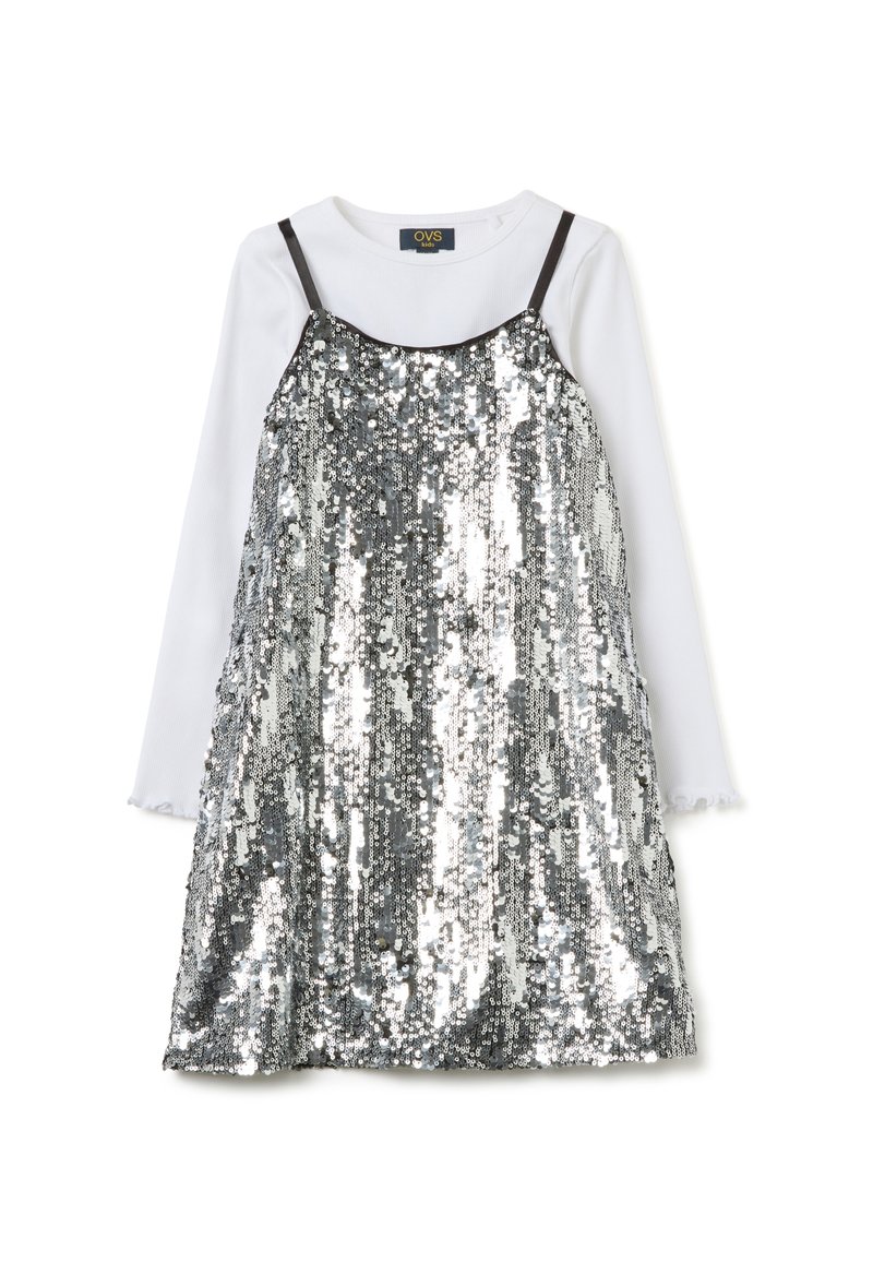 Vestido plateado con lentejuelas y tirantes oscuros ajustables, superpuesto sobre una camisa blanca de manga larga con puños con volantes, creando un efecto reflectante.