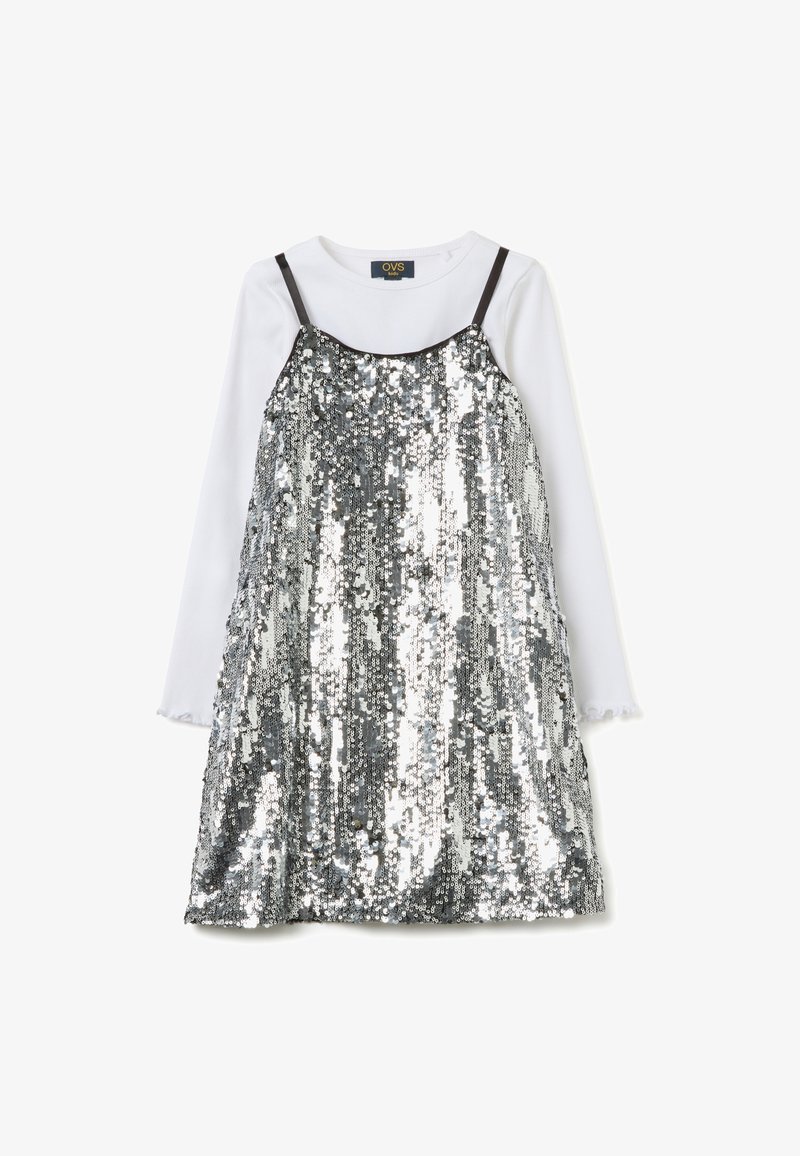 Vestido plateado con lentejuelas y tirantes oscuros ajustables, superpuesto sobre una camisa blanca de manga larga con puños con volantes, creando un efecto reflectante.