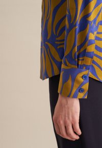 Camicia a maniche lunghe con un motivo astratto blu e senape, dotata di polsini con bottoni e una texture di tessuto liscia.