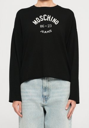 Czarny sweter z długim rękawem z białym napisem "MOSCHINO 86 - 23 JEANS" noszony z jasnoniebieskimi jeansami.