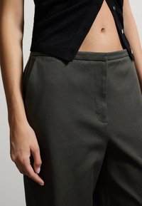 Pantaloni verdi scuri, sartoriali, con una trama liscia e tasche laterali. Accompagnati da un cardigan nero con bottoni. Dettaglio della vita visibile.