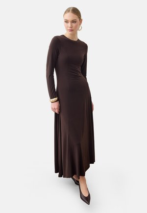 Femme portant une longue robe marron foncé cintrée avec des manches longues, des chaussures brunes pointues, des boucles d'oreilles créoles dorées et des bracelets dorés au poignet gauche.