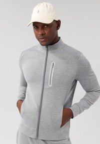 Šedá vetrovka na zips s vysokým golierom a prednou vreckom na zips, doplnená o svetlú čiapku. Hladký, priliehavý textil.
