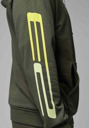 Mørkegrønn erme med et gult geometrisk stripemønster som går vertikalt langs armen, båret av en person mot en grå bakgrunn.