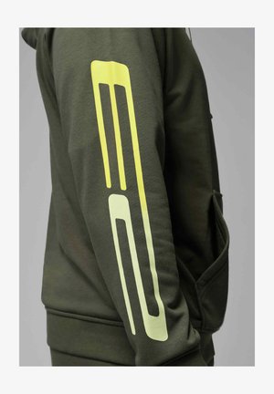 Mørkegrønn erme med et gult geometrisk stripemønster som går vertikalt langs armen, båret av en person mot en grå bakgrunn.