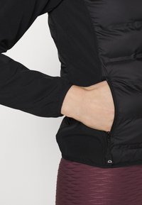 Chaqueta negra con una mezcla de tejido acolchado y suave, que presenta un bolsillo con cremallera y puños recogidos. Llevada con un fondo texturizado en color burdeos.