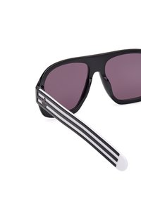 Ochelari de soare supradimensionați, negri, cu lentile de culoare violet închis, având o finisare mate și un braț lateral alb cu trei dungi negre.