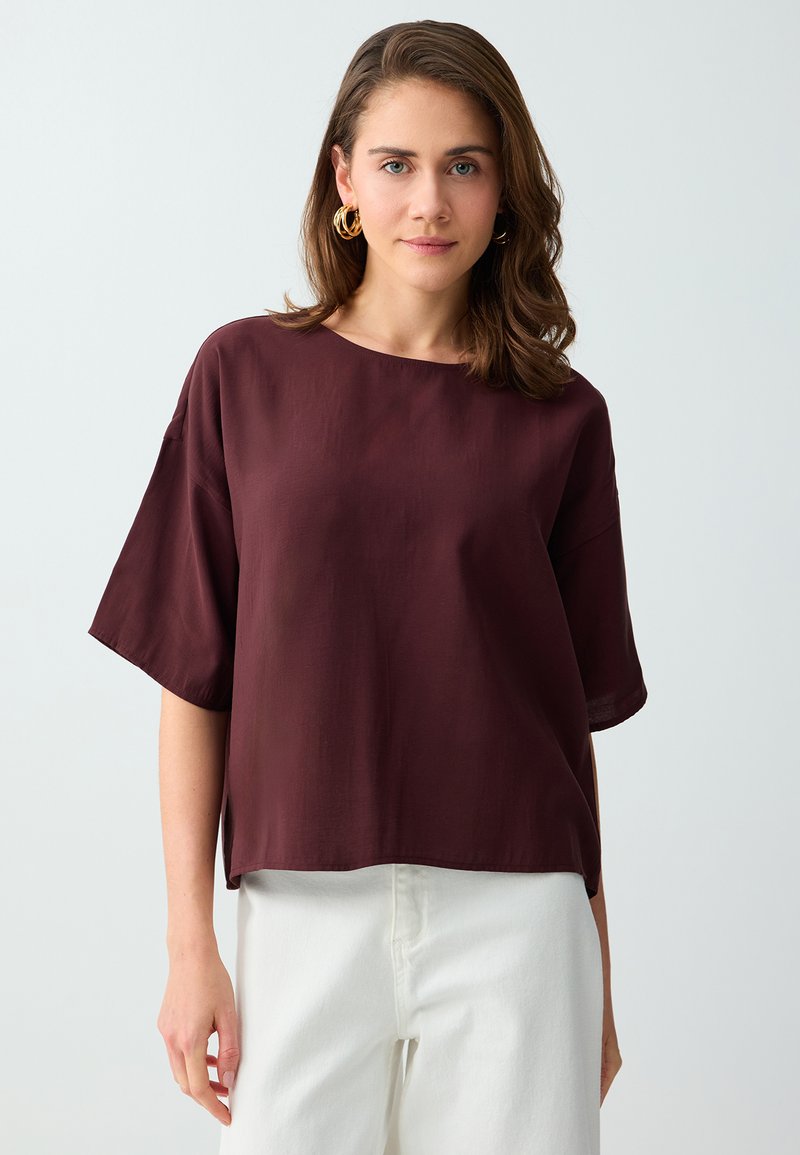 Jeune femme aux cheveux bruns mi-longs portant des boucles d'oreilles dorées en forme d’anneaux, un haut ample bordeaux foncé à manches courtes et un pantalon blanc, debout.