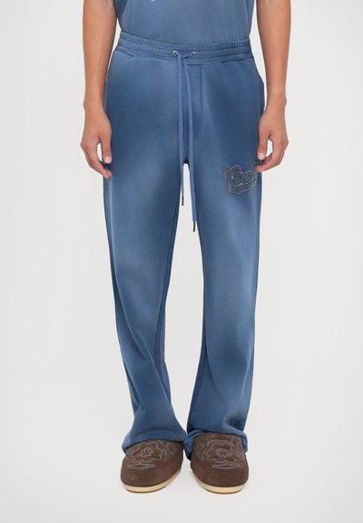 Mand iført blå afslappede bukser med snor i livet og brune broderede slip-on sko, stående mod en hvid baggrund.