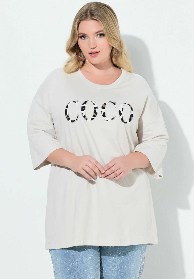 Lysebeige oversized t-shirt med rund halsudskæring, der har et sort-hvidt dyreprint med teksten 'COCO', kombineret med lyseblå glitrende jeans.