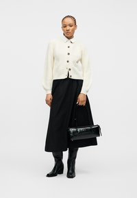 Γυναίκα που φοράει λευκό πουλόβερ με κουμπιά, μακριά μαύρη φούστα, μαύρες μπότες, κρατώντας μια μικρή μαύρη τσάντα χειρός σε απλό φόντο.