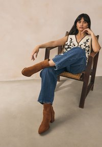 Bruine suède enkellaarsjes met een spits toelopende neus en een gestapelde hak, gecombineerd met blauw wijde pijpen jeans en een zwart-crème geborduurd vest.