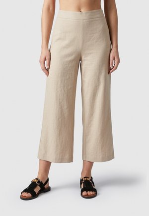 Beige wide-leg cropped pants worn with black open-toe sandals featuring gold buckle details on a person standing against a neutral background.