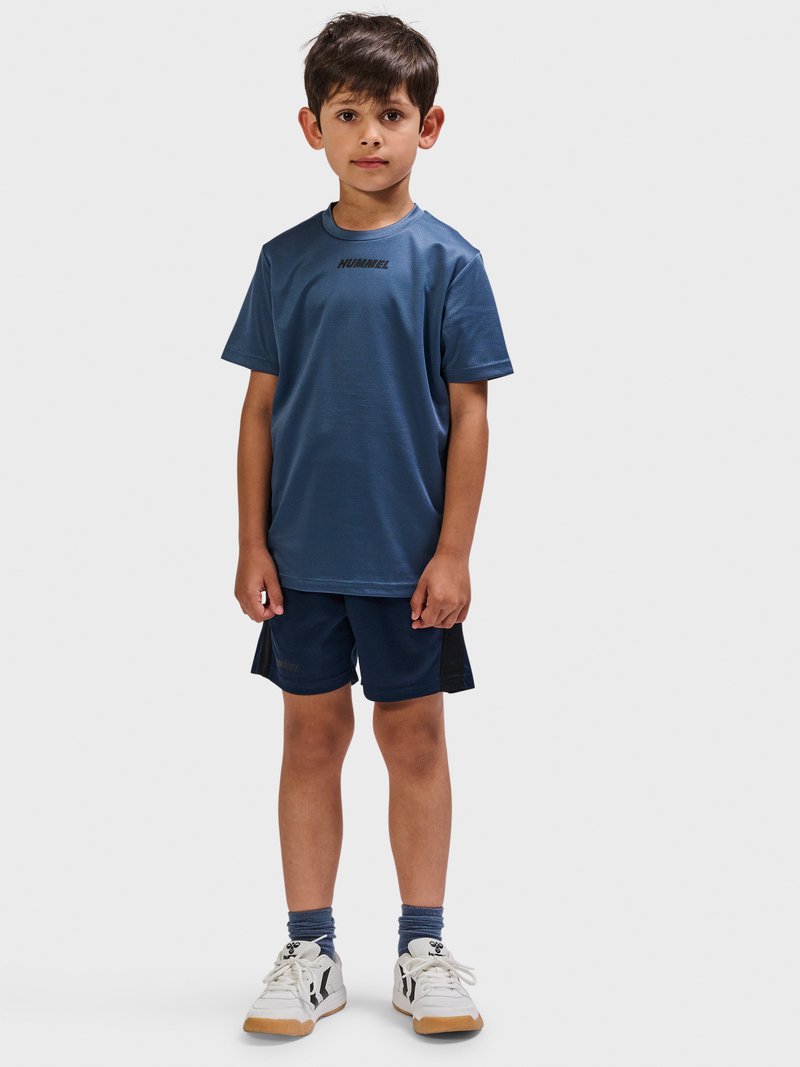 Junger Junge steht und trägt ein blaues Hummel-T-Shirt, passende Shorts, blaue Socken und weiße Sneakers vor neutralem Hintergrund.
