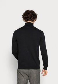 Homme aux cheveux bouclés portant un pull à col roulé noir et un pantalon à carreaux gris, debout de dos devant un fond uni clair.