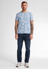 Světle modré tričko s bílým květinovým vzorem, krátké rukávy, spojené s tmavě modrými džíny a bílými teniskami. Muž s tetováním stojí vzpřímeně.
