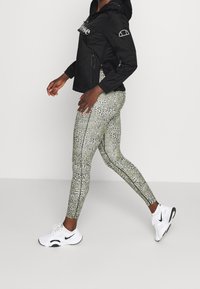 Sort letvægtsjakke med logo, kombineret med teksturerede grønne og sorte mønstrede leggings og hvide træningssko.