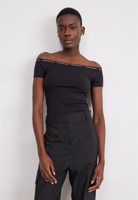 Černý top bez ramínek s logem Calvin Klein Jeans podél výstřihu, vyrobený z elastického materiálu; v kombinaci s černými kalhotami s vysokým pasem.