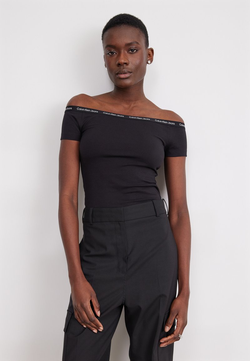 Černý top bez ramínek s logem Calvin Klein Jeans podél výstřihu, vyrobený z elastického materiálu; v kombinaci s černými kalhotami s vysokým pasem.