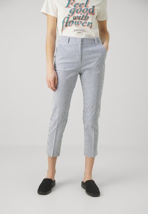Person iført blå og hvide stribede korte bukser, en hvid t-shirt med tekst og sorte slip-on sko stående mod en ensfarvet baggrund.