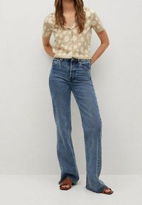 Beżowa koszula z krótkim rękawem w białe wzory stokrotek, zapinana na guziki; niebieskie, wysokie, rozkloszowane dżinsy z postrzępionym wykończeniem, zestawione z brązowymi sandałami.