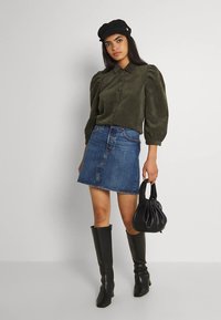 Donna che indossa una blusa verde oliva con maniche a sbuffo, una mini gonna di jeans, stivali neri fino al ginocchio, un berretto nero e tiene in mano una borsa nera.