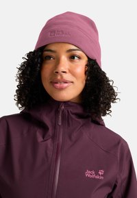 Vrouw met krullend haar, draagt een mauve fleece beanie en een bijpassende donkerpaarse jas met het Jack Wolfskin-logo, kijkt iets naar rechts.