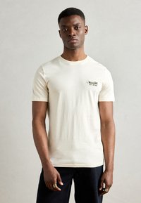 Tricou cu mânecă scurtă de culoare crem, din bumbac, cu guler rotund, având un design brodat verde și text în zona stângă a pieptului.