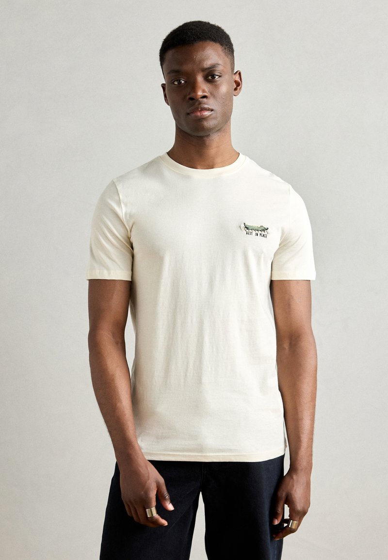 Tricou cu mânecă scurtă de culoare crem, din bumbac, cu guler rotund, având un design brodat verde și text în zona stângă a pieptului.