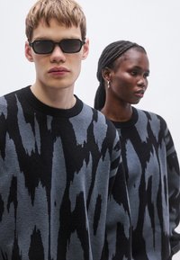 Črni in sivi puloverji z vzorcem in teksturirano površino. Ena modelka nosi sončna očala, medtem ko ima druga spete kite in uhani.