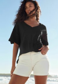 Zwarte V-hals T-shirt met opgerolde mouwen, gecombineerd met crèmekleurige shorts. De top heeft een relaxte pasvorm en een klein borstzakje.