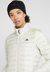 White puffer jacket with a high collar, featuring a black zipper and the Adidas logo. The model wears a ribbed black beanie with a red logo.