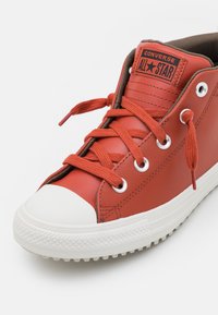 Czerwone skórzane trampki za kostkę z białą gumową podeszwą, metalowymi oczkami i pomarańczowymi sznurówkami; na języku widnieje logo 'Converse All Star'.