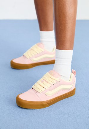 Rožiniai ir geltoni sportbačiai su nubuko viršumi, šviesiai geltonomis raištelėmis, guminiais padais ir šoninėmis juostelėmis. Dėvimi su baltais kojiniais.