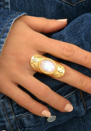 Bague en or avec une pierre de lune ovale au centre, gravures délicates autour de la monture, portée sur une main contre un tissu en denim.