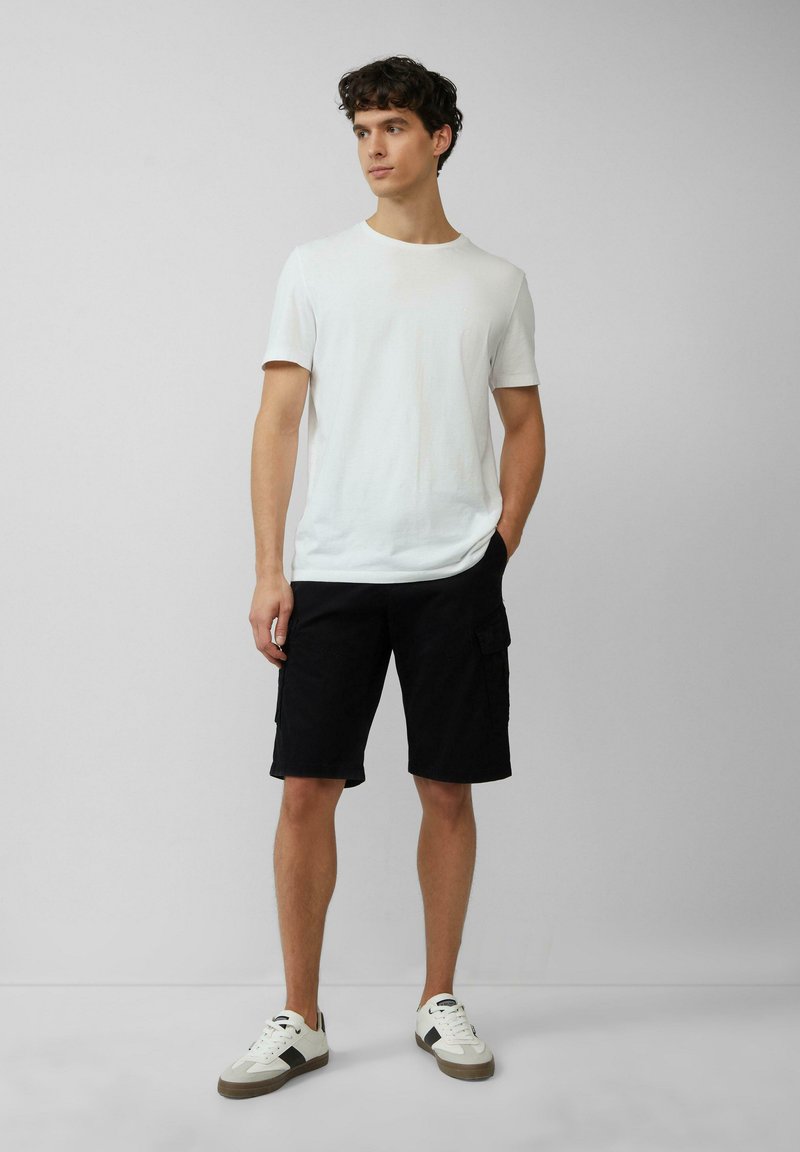 Witte katoenen t-shirt gecombineerd met knielange zwarte cargoshorts met zijzakken. De outfit wordt aangevuld met witte en grijze sneakers.