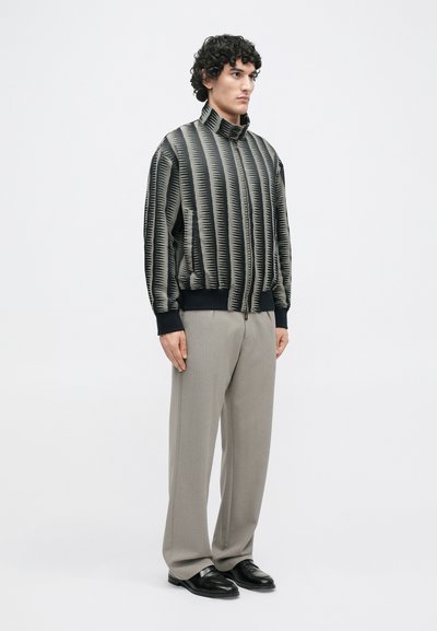 Bomberjacke in Schwarz und Beige mit vertikalen Streifenmustern, hoher Kragen und Reißverschluss. Kombiniert mit hellgrauer Hose und schwarzen Schuhen.