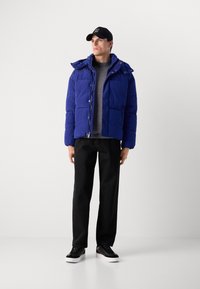 Blå pufferjakke med hette, teksturert stoff, frontlommer og trykkknapper, kombinert med sorte bukser og sorte joggesko.