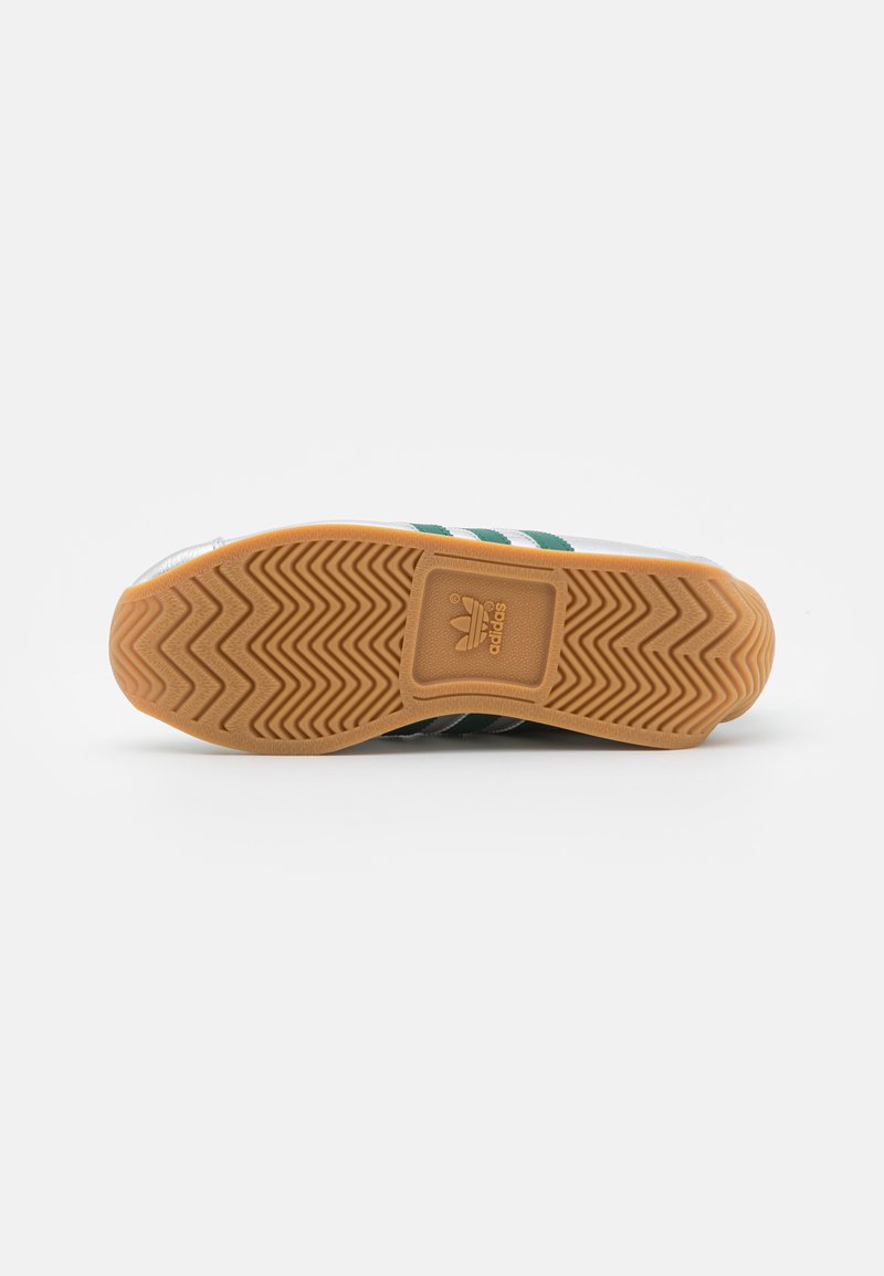 Białe sneakersy z zielonymi paskami, gumowa podeszwa z wzorem w jodełkę oraz logo na podeszwie. Prosty design, czyste linie.