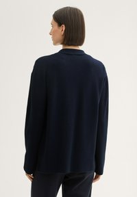 Cardigan tricoté bleu marine à coupe décontractée, avec manches longues, une texture douce et un ourlet fendu pour plus de liberté de mouvement.