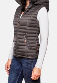 Mörkgrå vadderad väst med huva, utrustad med dragkedjefickor och en blank, quiltad yta. Bruna detaljer vid dragkedjorna.