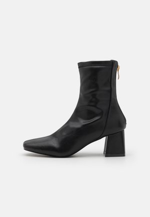 Botte en cuir noire mi-mollet avec bout carré, talon bloc et fermeture éclair arrière avec tirette dorée, vue de profil sur fond blanc.