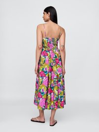 Frau mit langen dunklen Haaren, die ein ärmelloses, knielanges Blumenkleid mit rosa, grünen und blauen Blättern sowie flachen Sandalen trägt und von hinten schaut.