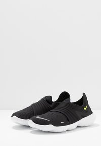 Czarne buty sportowe o smukłym wyglądzie, z teksturowaną cholewką z dzianiny, białą podeszwą i małym akcentem w neonowym żółtym kolorze.