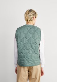 Gilet trapuntato in verde tenue con texture ondulata, caratterizzato da un colletto arrotondato e senza chiusura, indossato sopra una camicia bianca a maniche lunghe.