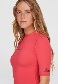 Červený krátky rukáv rash guard vyrobený zo syntetického materiálu, má okrúhly výstrih a logo na hrudi a rukáve. Hladká textúra.